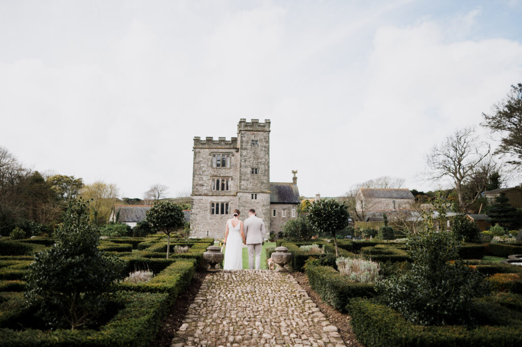 A newlywed couple walking towards a castle through the gardens of Pengersick Castle in Cornwall