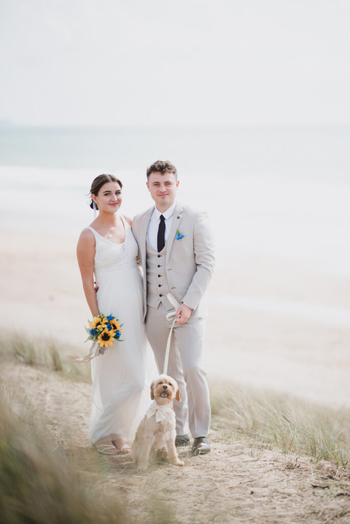 A newlywed couple and their dog standing on a bank leading down to the beach with the ocean in the background at Praa Sands in Cornwall