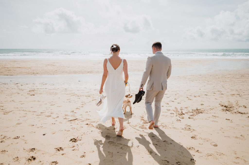 A newlywed couple and their dog walking towards the sea barefoot at Praa Sands in Cornwall