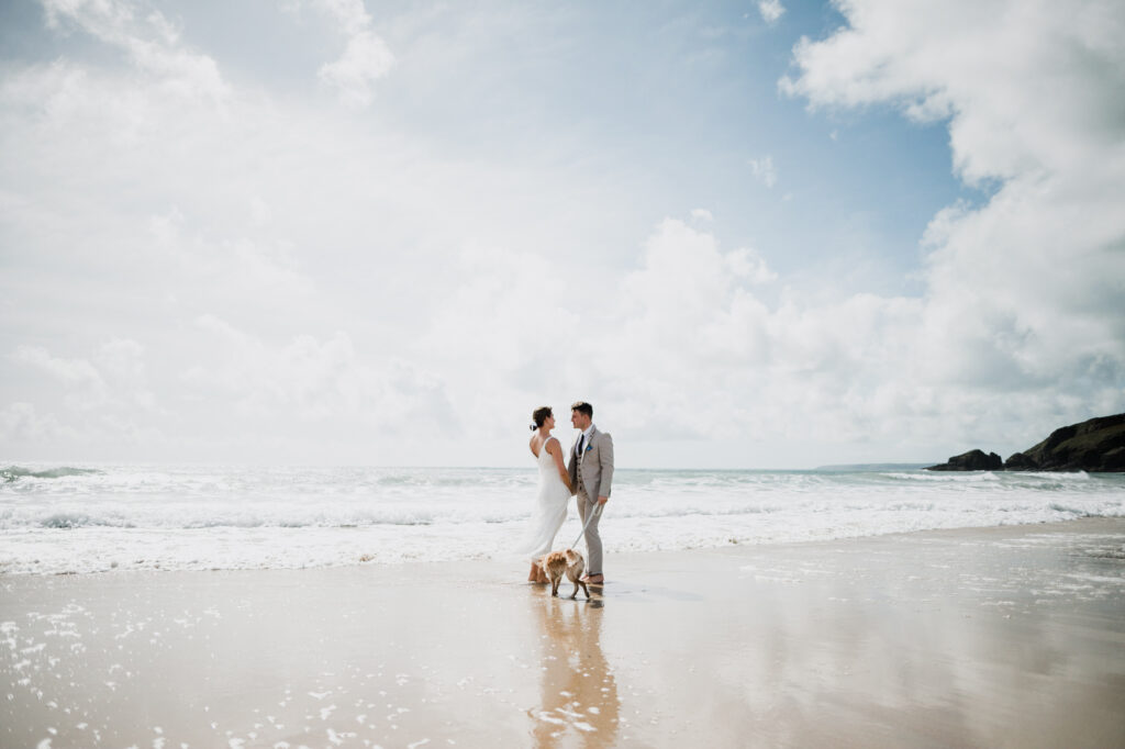 A newlywed couple standing close to the sea on the beach with waves breaking at their feet at Praa Sands in Cornwall