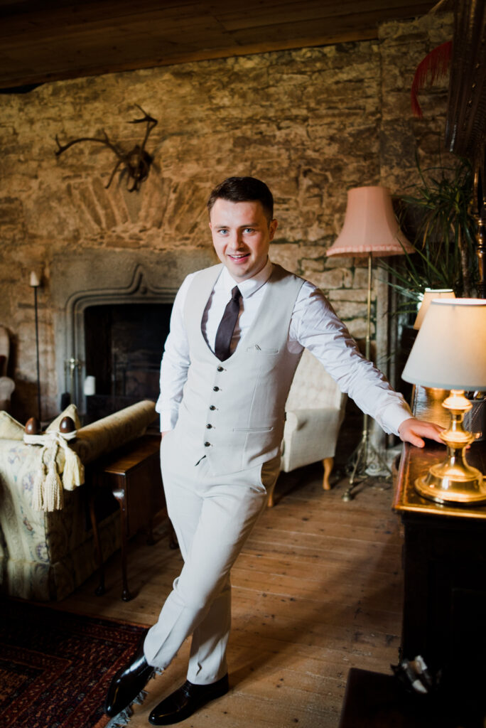A groom in his suit leaning on a dresser inside Pengersick Castle in Cornwall