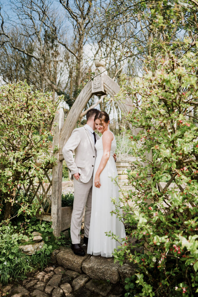 A newlywed couple standing in a wooden archway within the Pengersick Castle Gardens in Cornwall
